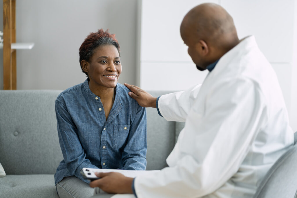 A male doctor gently placing a reassuring hand on the shoulder of a woman patient, who is smiling and seated on a couch. The interaction suggests a supportive and compassionate healthcare environment.